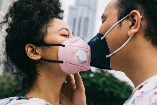 Young woman with frizzy black curly hair in a high ponytail is wearing a pink mask and kissing a man who is wearing a black mask and has his straight black hair in a bun.