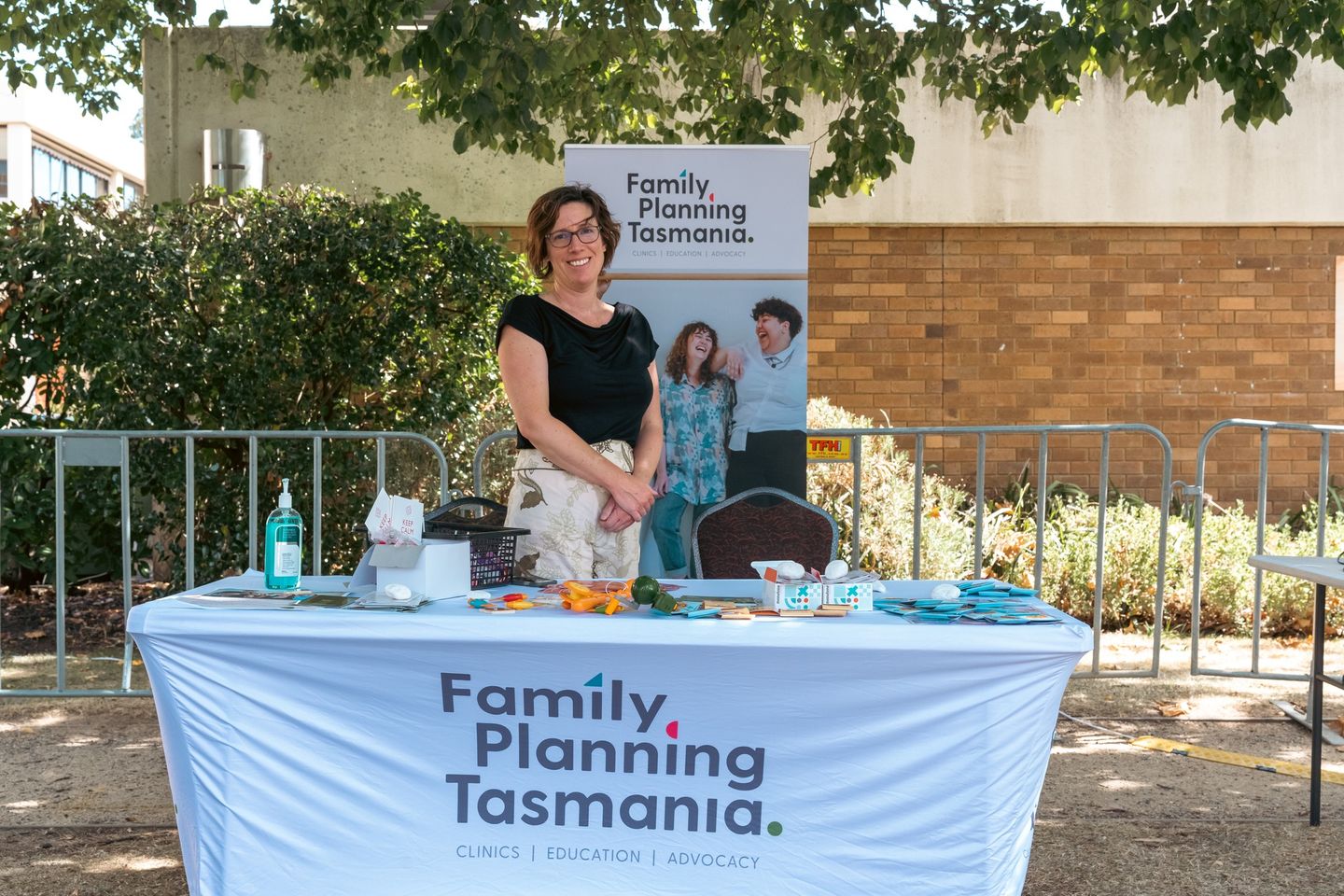 Clinical Nurse Alison smiling behind the table with a tablecloth that reads "Family Planning Tasmania" holding a stall promoting health at an event.
